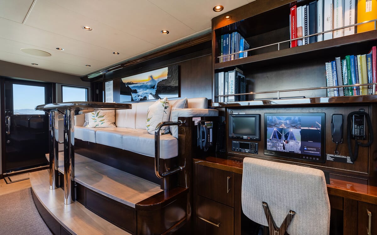 Interior view of the yacht FIGARO, featuring a lounge area with a sofa, bar table, and a desk area with monitor and a collection of books.