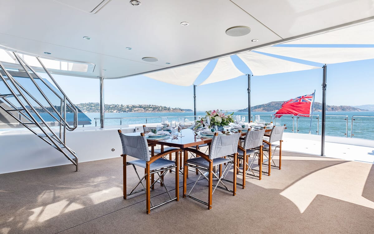 Exterior of the 39.62m yacht FIGARO. Dining table set for guests on the deck, British flag in the background, and sea view.