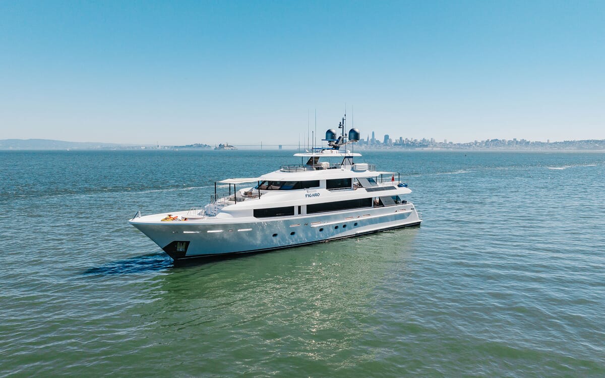 Aerial view of the 39.62m yacht FIGARO cruising on open water. The white yacht features multiple decks and is set against a city skyline.
