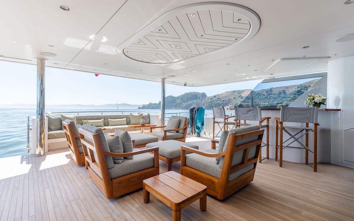 Exterior deck of the 39.62m yacht FIGARO. Comfortable seating area with sofas, armchairs and a small bar. The setting is at sea with coastline visible in the background.