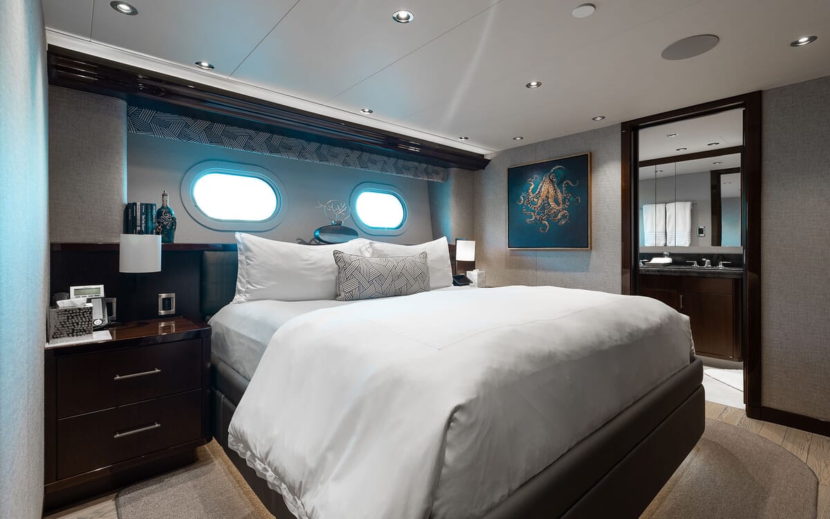 Interior view of the FIGARO yacht's master bedroom. The image shows a bed with porthole windows, artwork, and a view of the bathroom.