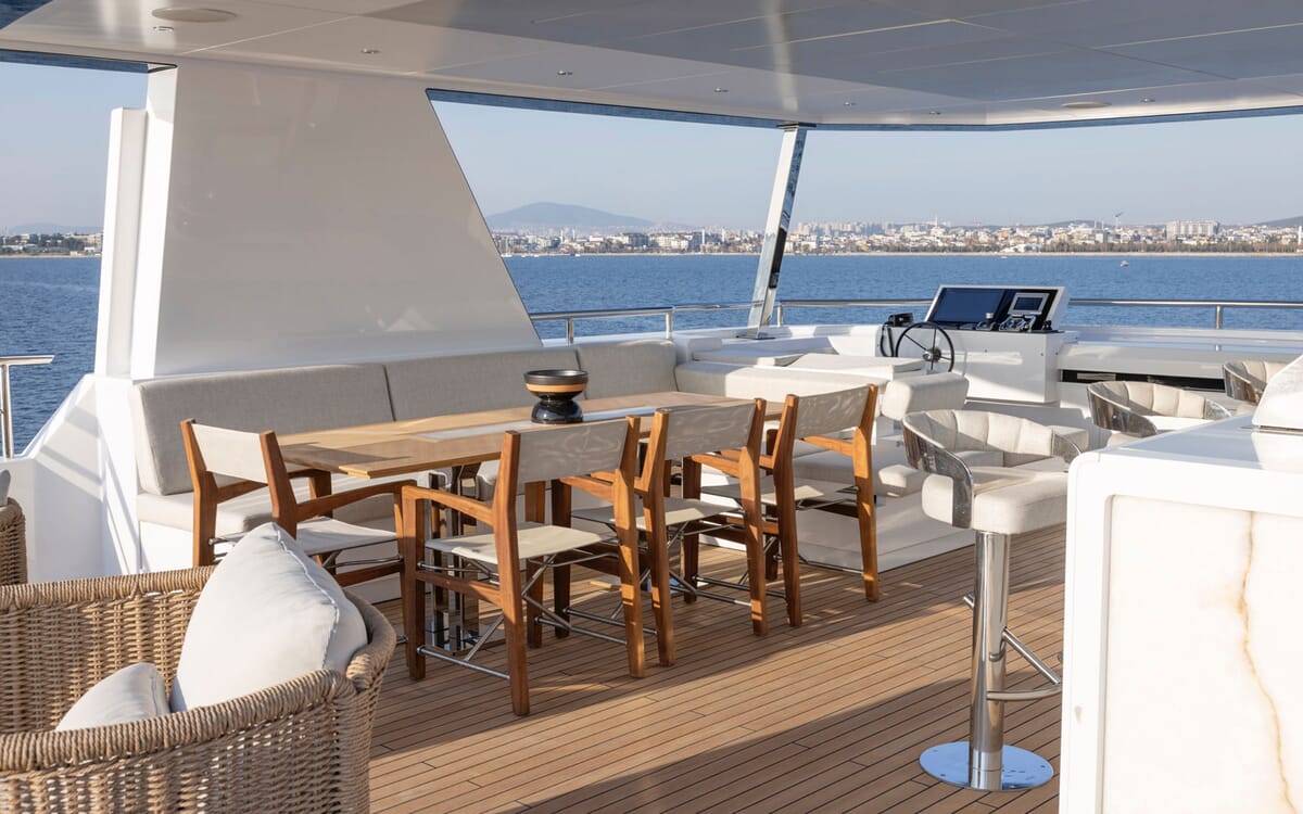 Vista exterior del BEE YACHT, un yate de 35,42 m. La imagen muestra una cubierta con asientos, una mesa y una zona de bar, con vistas al agua y a una ciudad.