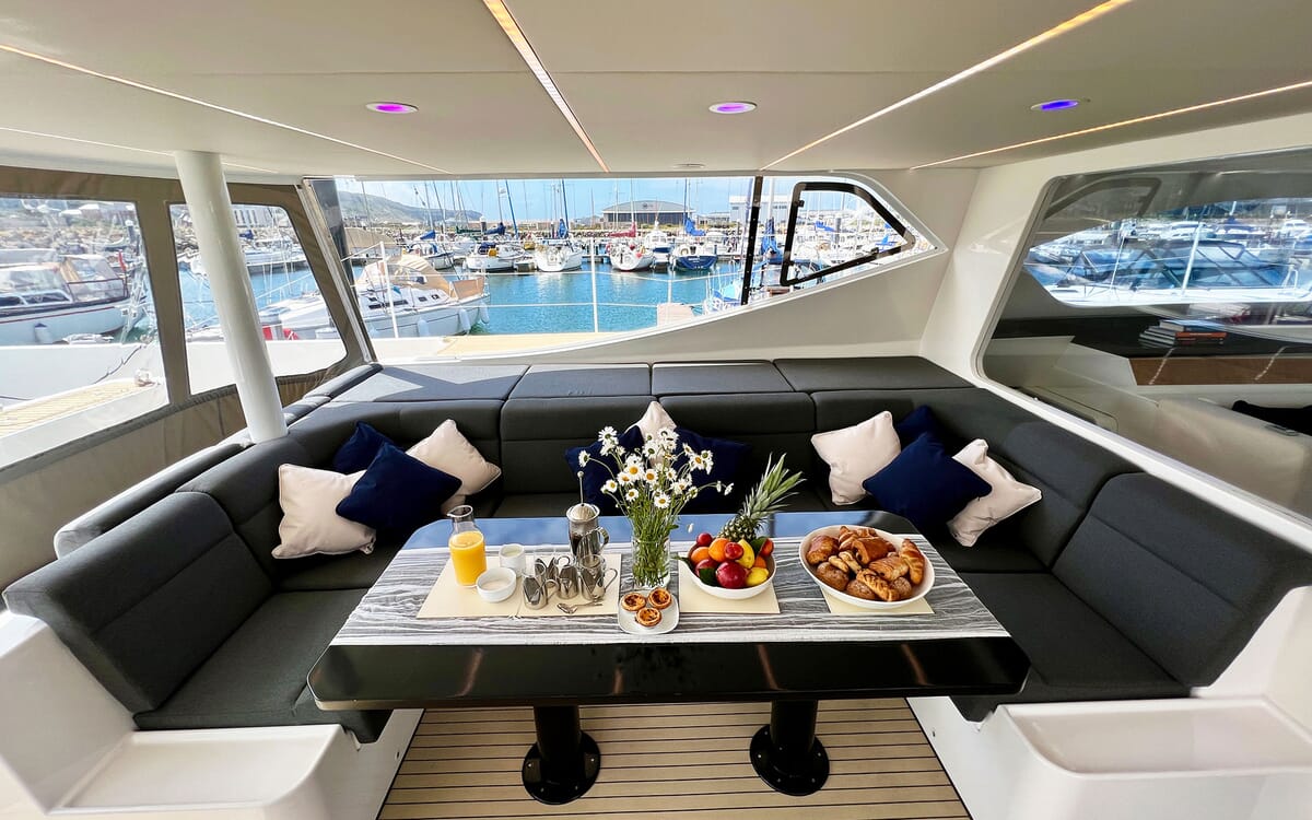Innenaufnahme der Yacht MAHAL KO (23,42 m LOA) mit einer Sitzecke und einem gedeckten Tisch für das Frühstück. Durch die Fenster ist ein Yachthafen zu sehen.