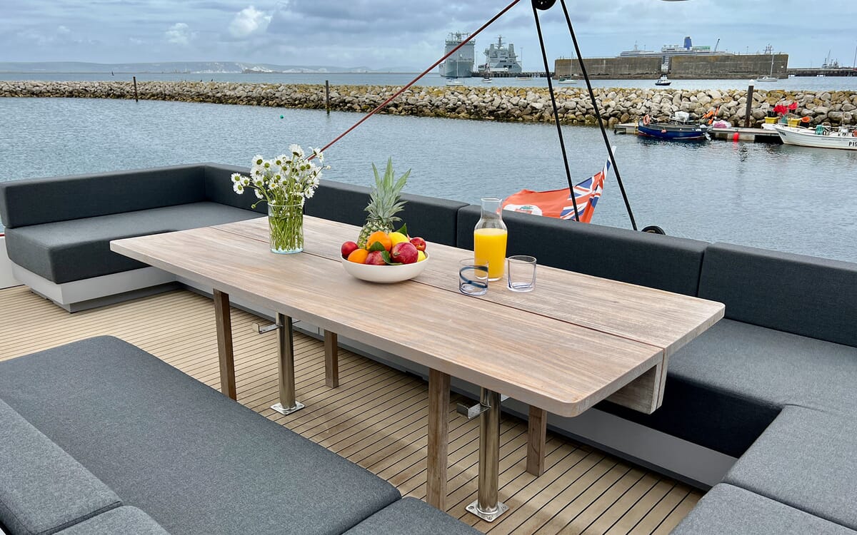 Außenansicht der Yacht MAHAL KO mit einem mit Obst und Blumen gedeckten Tisch. Die Yacht liegt vor Anker, im Hintergrund der Hafen.