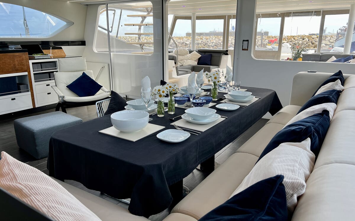 Innenansicht der MAHAL KO Yacht mit einem für eine Mahlzeit gedeckten Esstisch mit Blick auf einen Hafen. Zur Ausstattung gehören bequeme Sitzgelegenheiten und große Fenster.