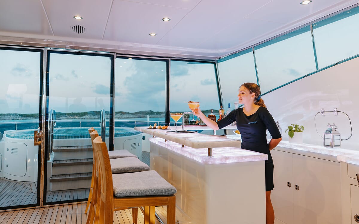 Im Inneren der 46,71 m langen Yacht ODYSSEA serviert ein Barkeeper Cocktails an einer beleuchteten Bar. Vor den großen Fenstern ist ein Jacuzzi zu sehen.