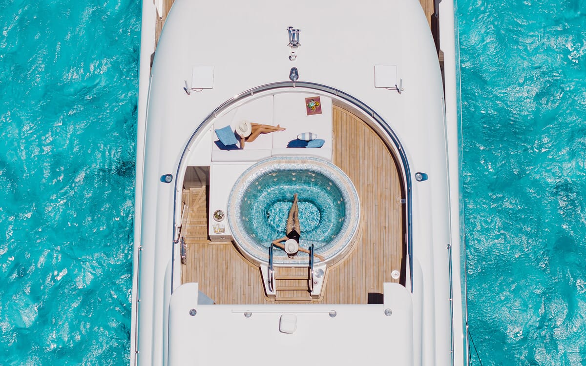 Luftaufnahme der ODYSSEA Yacht mit Sonnendeck, Jacuzzi und klarem, blauem Wasser. Zwei Personen entspannen sich an Deck.