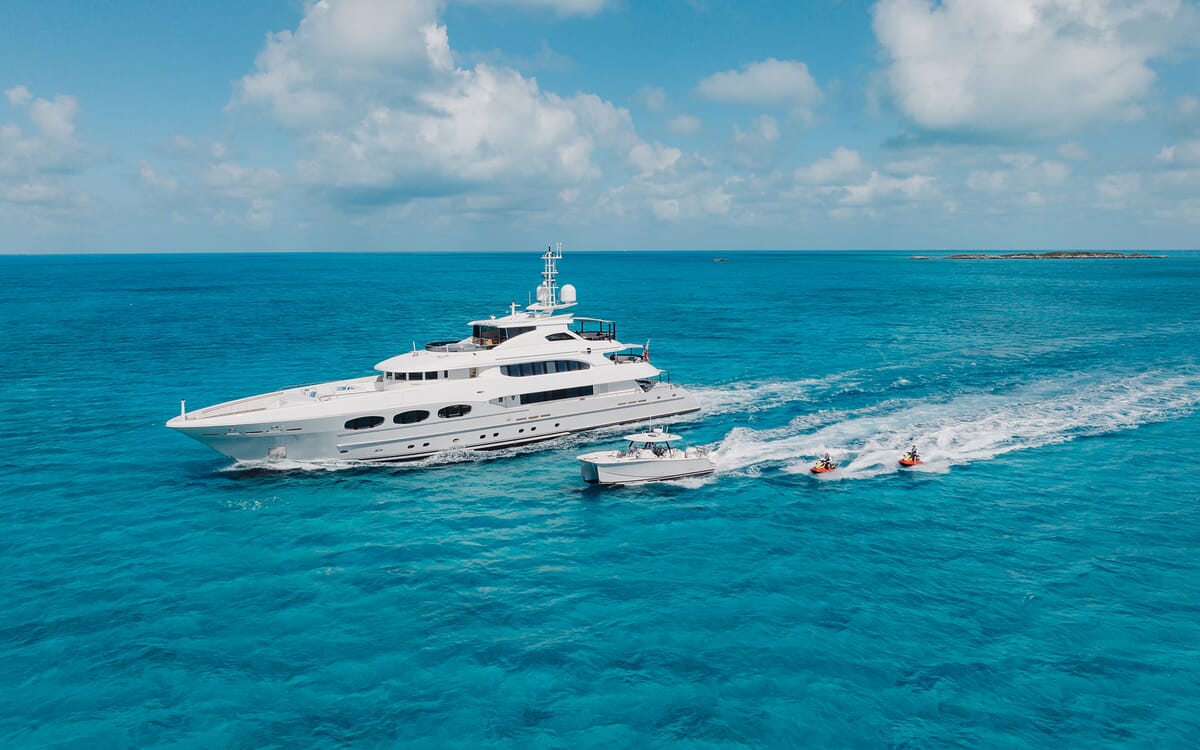 Luftaufnahme der 46,71 m langen Yacht ODYSSEA, die auf türkisfarbenem Wasser unterwegs ist. Unter strahlend blauem Himmel sind auch ein Beiboot und Jetskis zu sehen.