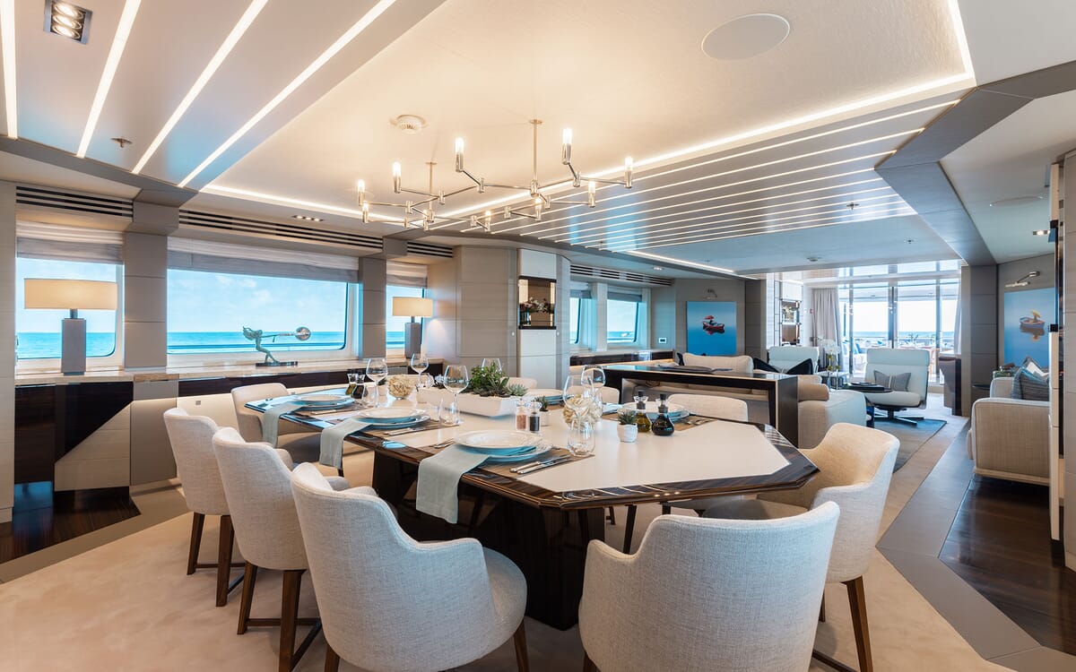 Innenansicht des Essbereichs der ODYSSEA Yacht mit einem großen Tisch, der für eine Mahlzeit gedeckt ist, heller Beleuchtung und einem malerischen Meerblick.